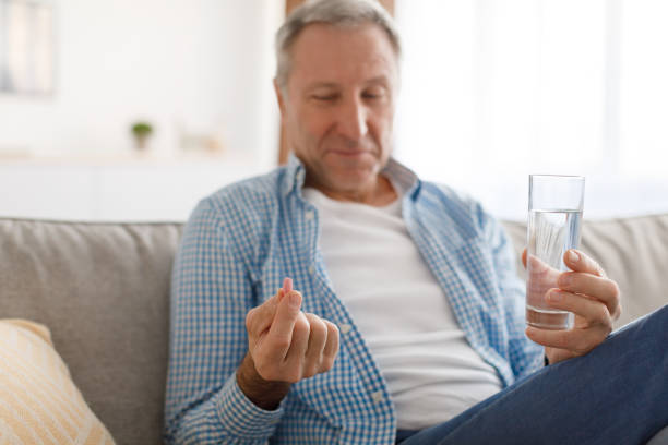 Closeup of senior man sitting on couch with glass of water, holding pill in hand taking painkillers at home. Mature male patient using medications, selective focus, blurred background|smiling senior couple with pill bottle sitting in sofa at home||