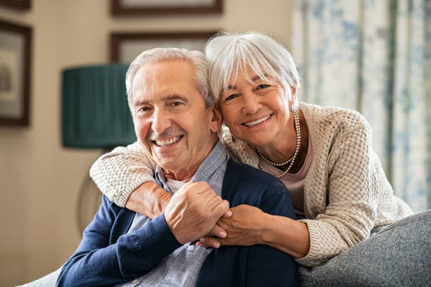 Portrait of romantic senior man with his beautiful wife stay at home. Smiling and caring old woman embracing from behind her retired husband sitting on couch. Cheerful old couple looking at camera with joy.|||